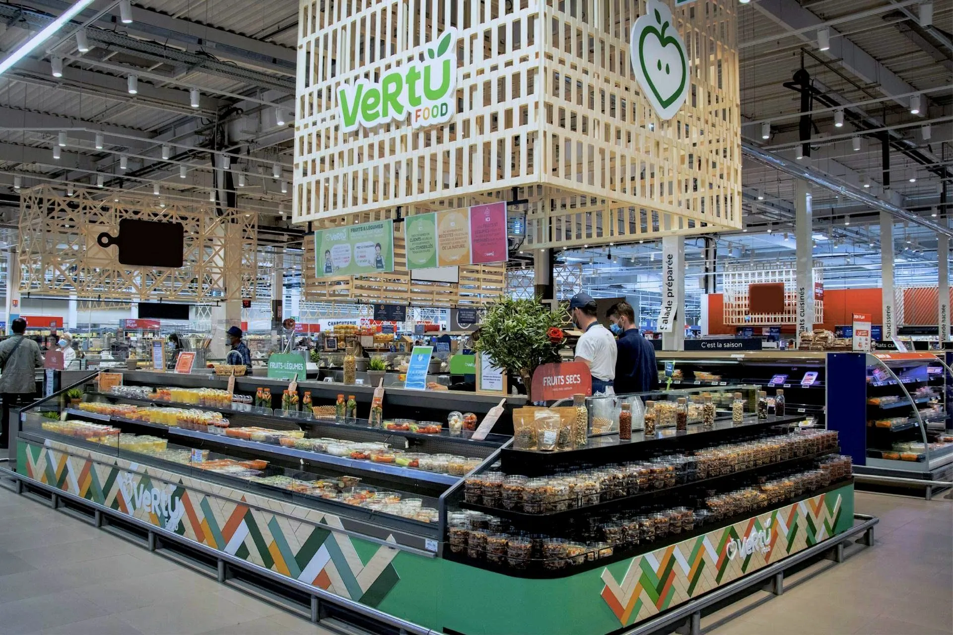Kiosque Vertu Food en hypermarché : enseigne en lattes de bois, comptoirs à chevrons colorés, fruits secs et salades